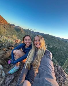 two women sitting on top of a mountain with their arms around each other and smiling at the camera