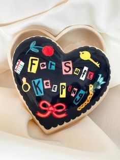 a black heart shaped box with letters and scissors on it's side sitting on a white cloth