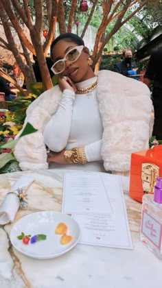 a woman sitting at a table wearing glasses and a fur coat over her shoulders, with food on the table in front of her