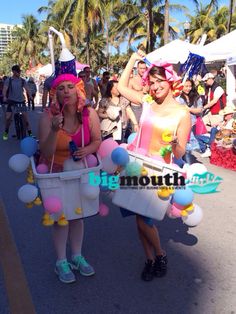 two women in costumes are walking down the street with buckets on their heads and balloons around their necks