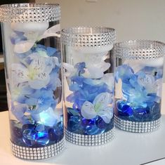 three glass vases with blue and white flowers in them on a counter top next to each other