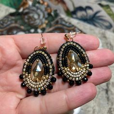 ✨ Elevate your elegance with these Black and Gold handmade earrings, crafted with meticulous attention by me. At the heart of each earring, a radiant high-quality Golden crystal takes center stage, surrounded by a symphony of matte and shiny black seed beads. The crystal is adorned with opulence, featuring accents of 24K Gold Delica Beads and matt silver seed beads, all gracefully wrapped in a rhinestone chain. 🌟 Dangling from your ears with grace, these earrings are secured with an 18K gold ballpost earwire, ensuring a secure and stylish fit. The dark gray faux leather backing adds a touch of sophistication, making these earrings a versatile accessory for any occasion. ✨ **Materials - Gold Crystal: A center of attention, radiating timeless beauty. - Seed Beads: A harmonious blend of matt Black Beaded Earrings Native American, Traditional Black Beaded Earrings With Fringe, Black And Gold Beaded Earrings, Traditional Black Beaded Fringe Earrings, Elegant Black Handwoven Beaded Earrings, Black And Gold Beaded Drop Earrings, Classy Earrings, Gold Bead Earrings, Native American Beaded Earrings