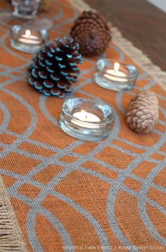 pine cones and candles are sitting on a placemat with an orange table runner in the background