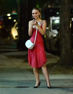 a woman in a red dress is holding a white purse and looking at her cell phone