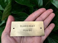 a hand holding a brass plaque that says claremont house in black lettering on it, surrounded by green leaves