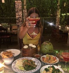 a woman holding up a wine glass in front of her face while sitting at a table filled with food