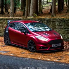 a red car parked on the side of a road next to a stone wall and trees