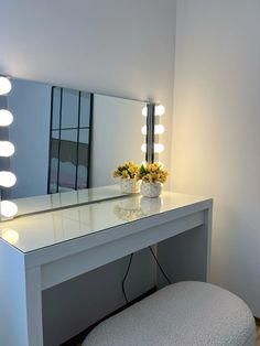 a white vanity with flowers and lights on the top shelf next to a stool in front of a mirror