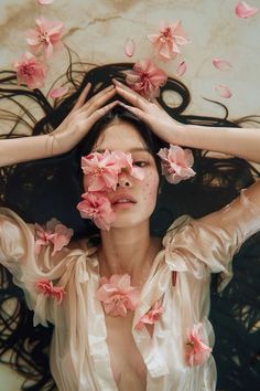 a woman with flowers in her hair covering her eyes and hands behind her head, surrounded by pink petals