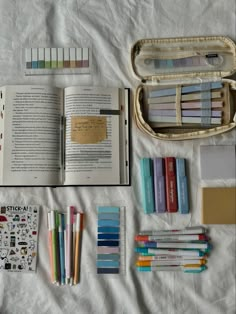 an open book sitting on top of a bed next to books and pencils in a bag