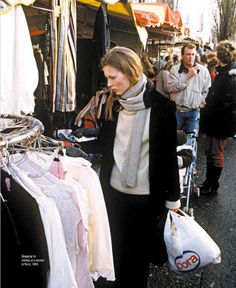 a woman standing in front of a market with clothes on display and people walking by