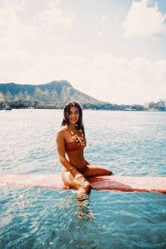 a woman sitting on top of a surfboard in the middle of the ocean with mountains in the background