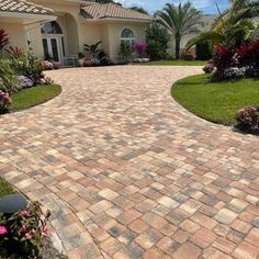 a driveway in front of a house with flowers on the side and palm trees around it