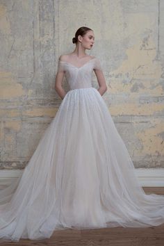 a woman standing in front of a wall wearing a wedding dress with an off the shoulder neckline