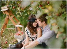 a man and woman sitting on the ground in front of an apple tree holding each other