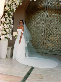 a woman in a wedding dress standing next to a flower covered archway with white flowers