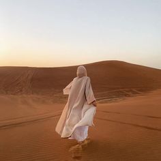 a person walking in the desert wearing a white robe and headdress, with sand dunes in the background