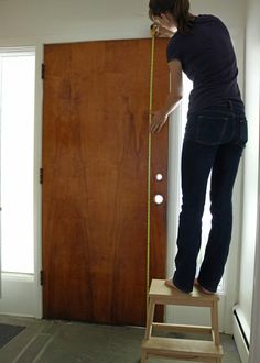 a woman standing on a step stool measuring the length of a wooden door with a tape measure