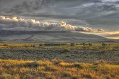 an open field with mountains in the distance and clouds above it on a cloudy day