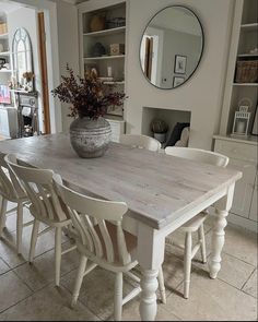 a dining room table with white chairs and a vase on top of it in front of a mirror