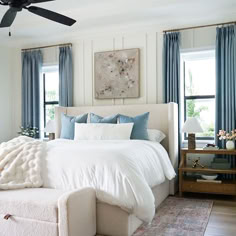 a white bed sitting in a bedroom next to two windows with blue drapes on them