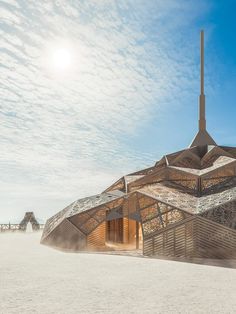 an unusual building sits on the beach with sand blowing in front of it and blue skies above