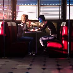 two people sitting at a table in a diner with red booths and windows behind them