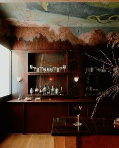 an empty bar with many bottles and glasses on the counter, in front of a painting