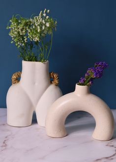 two white vases with flowers in them sitting on a marble countertop against a blue wall