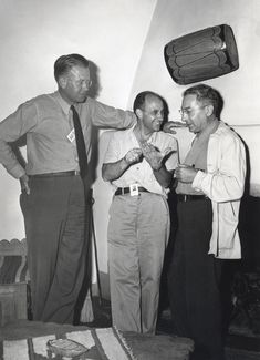 black and white photograph of three men standing in front of a television set with one man pointing at another man's tie