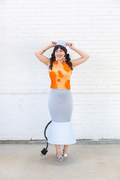 a woman in an orange top and grey skirt with her hands on her head while standing next to a white brick wall