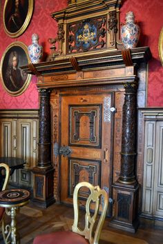 an old wooden cabinet with paintings on the wall behind it and two chairs in front of it