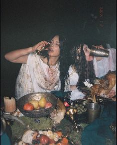 two women sitting at a table with food and drinks in front of them, drinking from cups