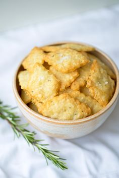 a bowl filled with crackers next to a sprig of rosemary on a white cloth