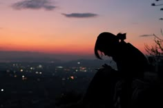 a woman sitting on top of a hill next to a city at night with the sun setting