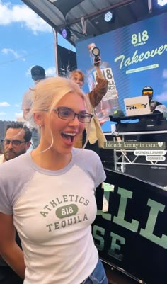 a blonde woman with glasses standing in front of a sign at an outdoor event and smiling