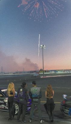 four people standing in front of a race car watching fireworks go off over the track