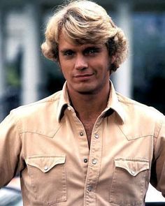 a man with blonde hair wearing a tan shirt and looking at the camera while standing in front of a car