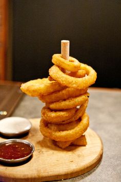 a stack of onion rings sitting on top of a wooden board next to dipping sauces