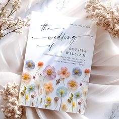 a wedding card with flowers on it sitting next to some white fabric and dried grass