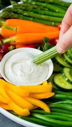 a person dipping dip into a bowl of veggies on a platter with asparagus, carrots and cucumbers