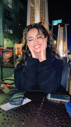 a woman sitting at a table smiling and holding her hand to her face, in front of a cityscape