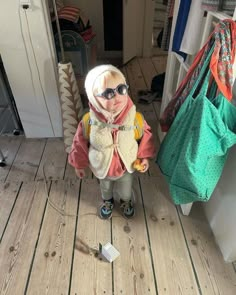 a little boy wearing sunglasses and a scarf standing in front of a door with bags on the floor