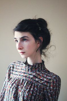 a young woman with black hair wearing a blouse and earrings, standing in front of a white wall