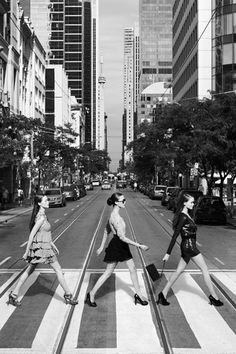 three women crossing the street in front of tall buildings on a crosswalk with their legs crossed