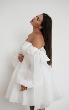 a woman with long hair wearing a white off the shoulder dress and holding her hands behind her back