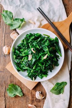 a white bowl filled with green vegetables on top of a wooden table next to chopsticks