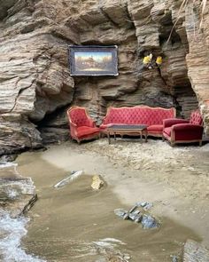 two red couches sitting on top of a sandy beach next to a cliff face