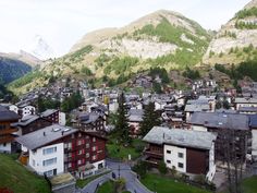 a town with mountains in the background and houses on the hill side, surrounded by green grass