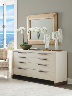 a white dresser with flowers and vases on it in front of a large window